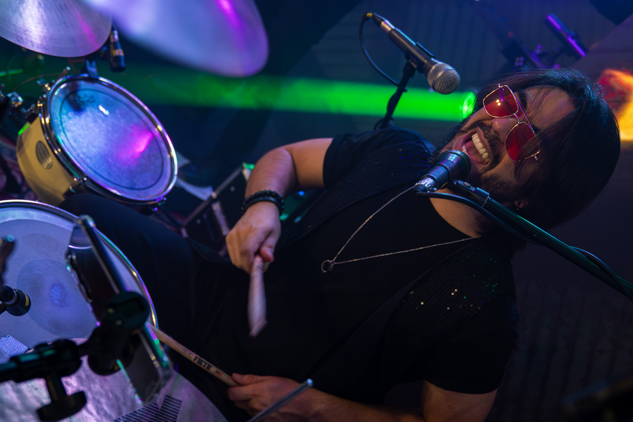 Músico dos Os Manteigas cantando e tocando bateria, com óculos vermelhos e iluminação colorida.